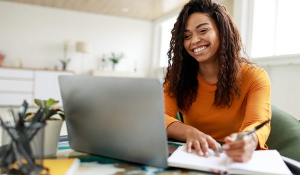 Distance Education. Portrait of smiling woman sitting at desk, using laptop and writing in notebook, taking notes, watching tutorial, lecture or webinar, studying online at home looking at screen